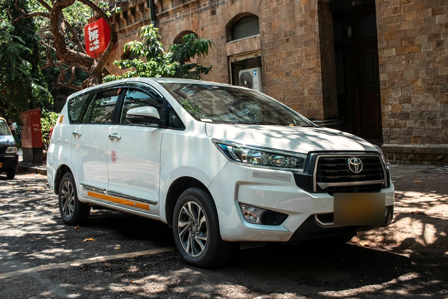 White Toyota Innova Crysta parked in dappled shade beside an old stone building